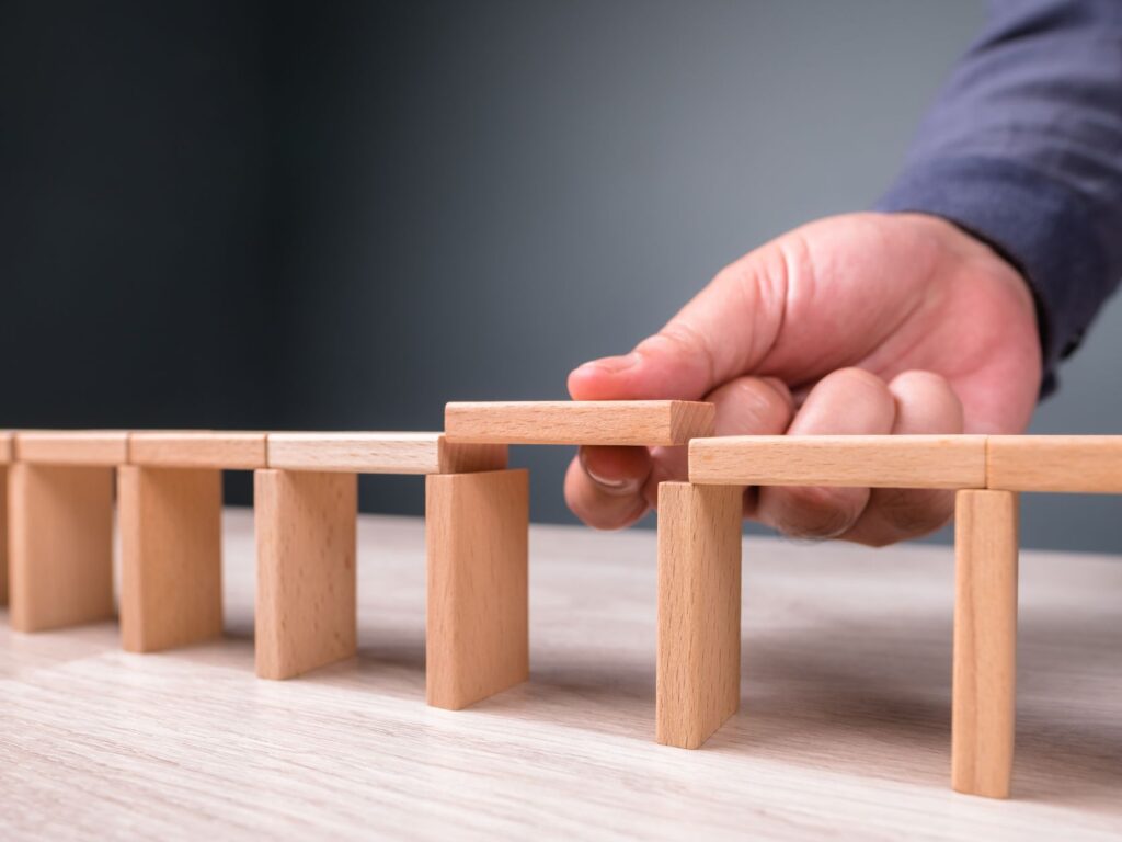 A hand places the final wooden block to connect two sides of a small bridge made of wooden pieces.