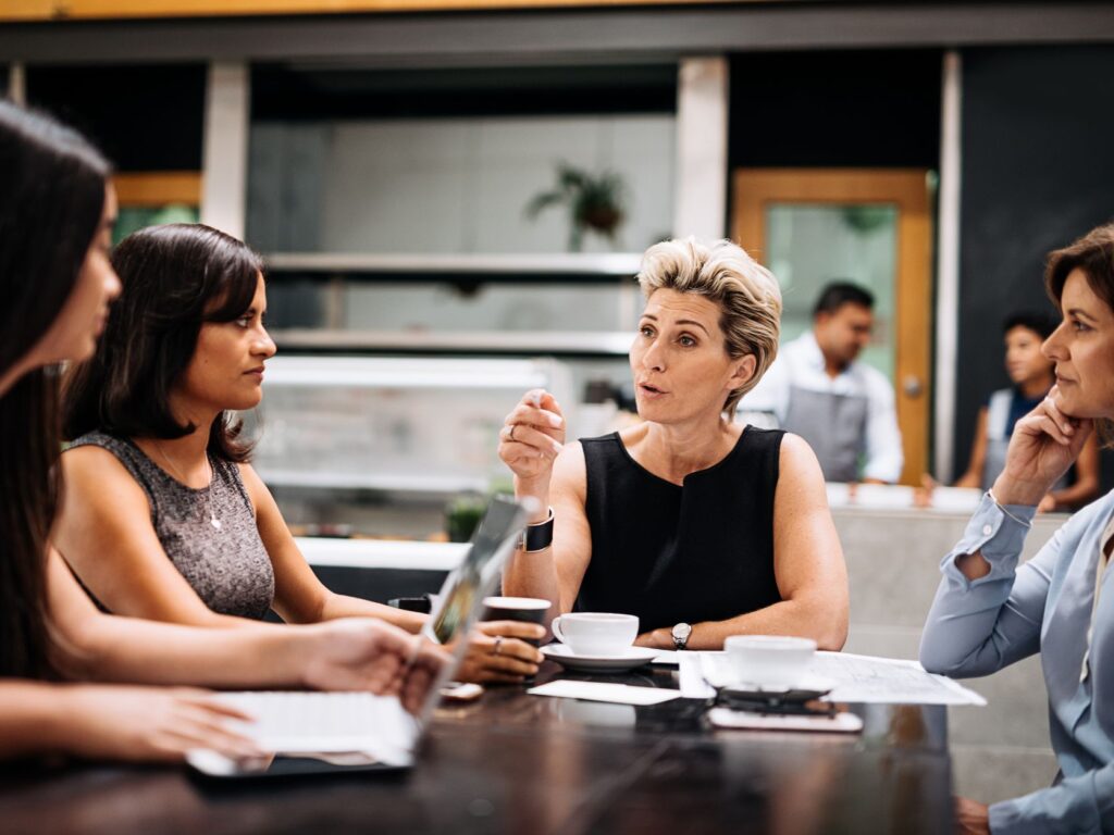 Quatro mulheres reunidas à mesa de um café/escritório; uma delas fala enquanto as outras escutam, com laptop, papéis e xícaras à vista.