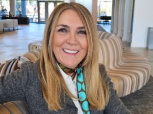 Smiling blonde woman wearing a gray blazer and patterned scarf, seated in a bright, modern indoor space with contemporary lounge chairs in the background.