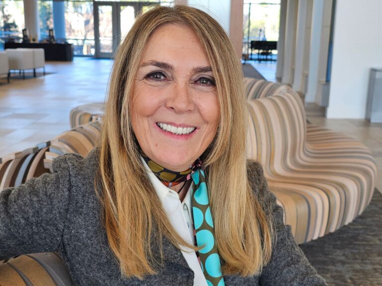 Smiling blonde woman wearing a gray blazer and patterned scarf, seated in a bright, modern indoor space with contemporary lounge chairs in the background.