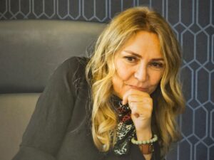 A middle-aged woman with long blonde hair sits indoors in front of large window panels overlooking a courtyard with a fountain. She wears a beige sweater vest over a white shirt and a pendant necklace, creating a warm and approachable professional portrait in a modern office setting.