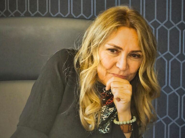 A middle-aged woman with long blonde hair sits indoors in front of large window panels overlooking a courtyard with a fountain. She wears a beige sweater vest over a white shirt and a pendant necklace, creating a warm and approachable professional portrait in a modern office setting.