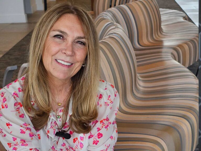 Smiling woman with long light brown hair wearing a floral blouse, seated indoors beside striped upholstered chairs.