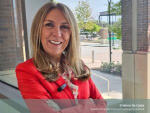 Smiling woman in a red blazer seated indoors by a large window, with on-screen text identifying her as Cristina Da Costa, Coach for Leadership and Confidence at Work.