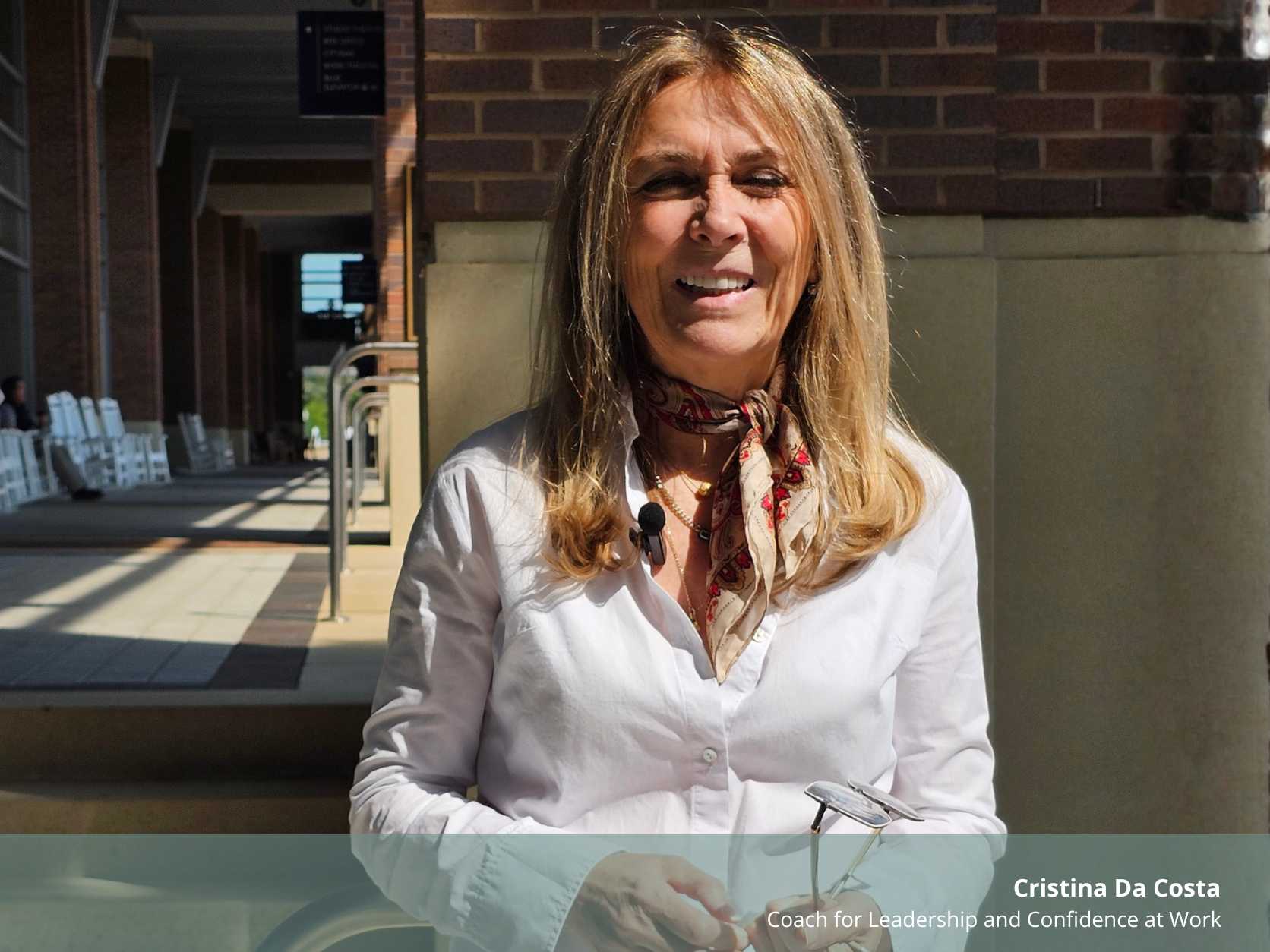 Retrato de Cristina Da Costa sorrindo ao ar livre. Ela é uma mulher de meia-idade, com cabelo loiro comprido, veste uma camisa branca e um lenço estampado ao pescoço. No canto inferior direito, lê-se o texto informativo com o seu nome e cargo profissional.