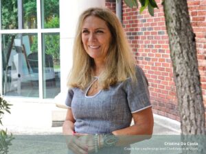 A professional, warm portrait of smiling Coach Cristina Da Costa, identified by text on the image as "Cristina Da Costa, Coach for Leadership and Confidence at Work." She is standing outdoors with clasped hands, between a brick wall and a glass window. The file is named 'The Question That Cuts Through the Noise Cristina da Costa.jpg'.