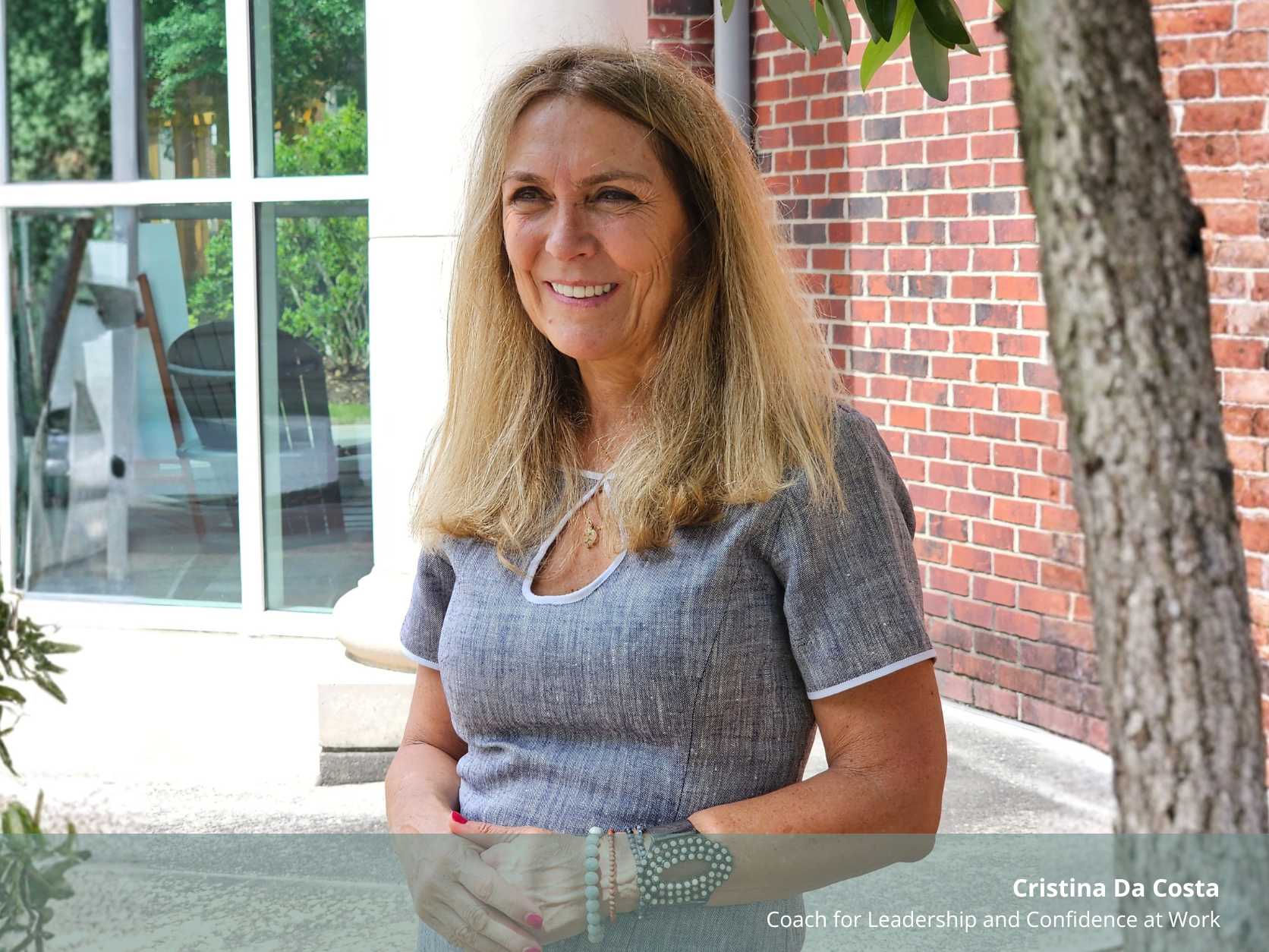 A professional, warm portrait of smiling Coach Cristina Da Costa, identified by text on the image as "Cristina Da Costa, Coach for Leadership and Confidence at Work." She is standing outdoors with clasped hands, between a brick wall and a glass window. The file is named 'The Question That Cuts Through the Noise Cristina da Costa.jpg'.